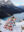 Person standing on a snowy mountain overlook holding a quilted blanket decorated with illustrated Canadian wildlife, landmarks, and cultural symbols above a turquoise alpine lake.