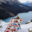 Person standing on a snowy mountain overlook holding a quilted blanket decorated with illustrated Canadian wildlife, landmarks, and cultural symbols above a turquoise alpine lake.