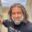 Portrait of photographer Greg Gerla with long gray hair and a beard, smiling outdoors between weathered concrete walls with a softly blurred landscape in the background.