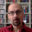 Portrait of artist Misa Nikolic wearing round glasses and a red collared shirt, seated in front of bookshelves filled with books and CDs.