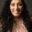 Portrait of designer Vishu Mahajan smiling at the camera, with long dark curly hair, wearing a light pink lace blouse, against a dark studio background.