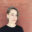 Portrait of Nicole Burisch standing in front of a red brick wall, wearing a black shirt and teal circular earrings.