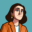 Illustrated self-portrait of AUArts alumna Ashrang Mohammadakbar featuring a stylized character with short brown hair, an orange jacket, and a teal background.
