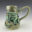 Hand-built stoneware beer stein by Gillian Boon with green and blue glazes, layered sculptural forms and small metal charms suspended from its large curved handle.